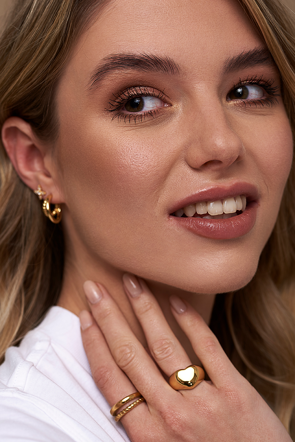 Model on a beige background, showcasing earrings and rings. Having her fingers placed on her chin so you can see the rings easy.