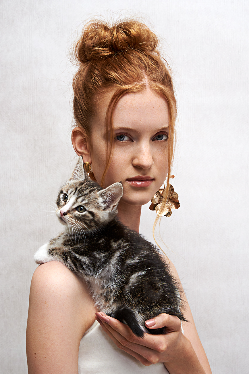 Model on a grey textured background holding a kitten in her hand which lies on her shoulder.