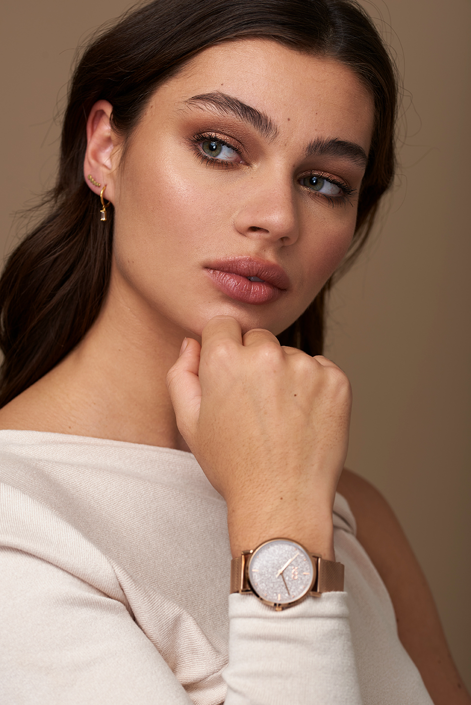 Model on a beige background with darker hair, holding her hand close to her face to showcase her watch.