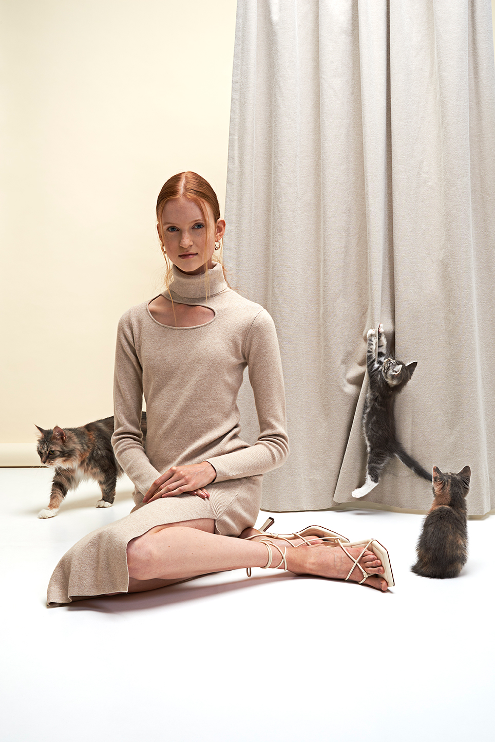 Model sitting in front of a curtain while kittens are climbing in the curtains.