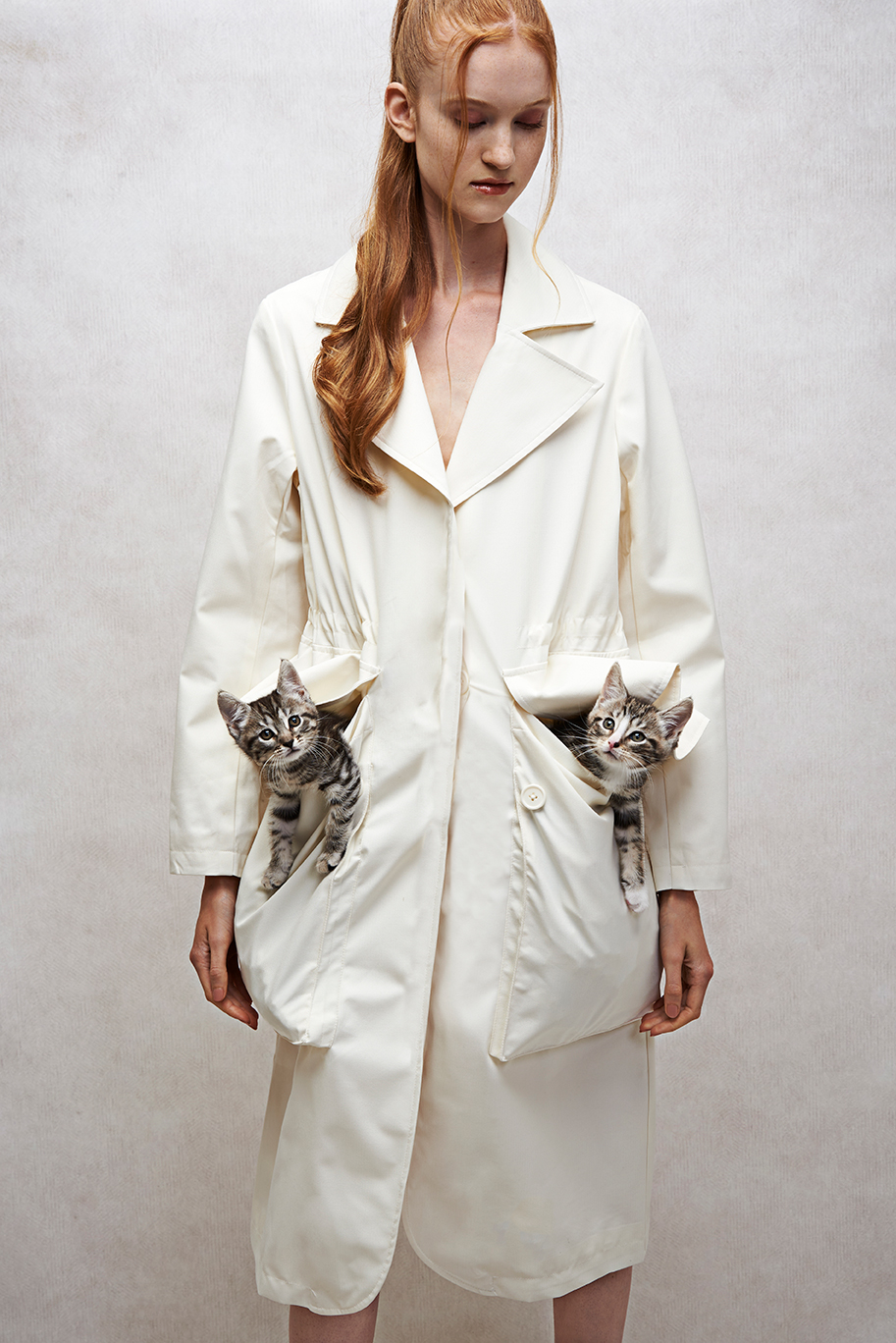 Model on a grey textured background wearing a jacket with two kittens in her pockets.